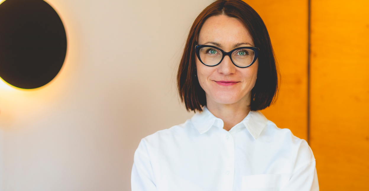 Na zdjęciu uśmiechnięta Anna Sarnacka-Smith ubrana w białą koszulę. W tle widoczna szafa i jasny kolor ściany.